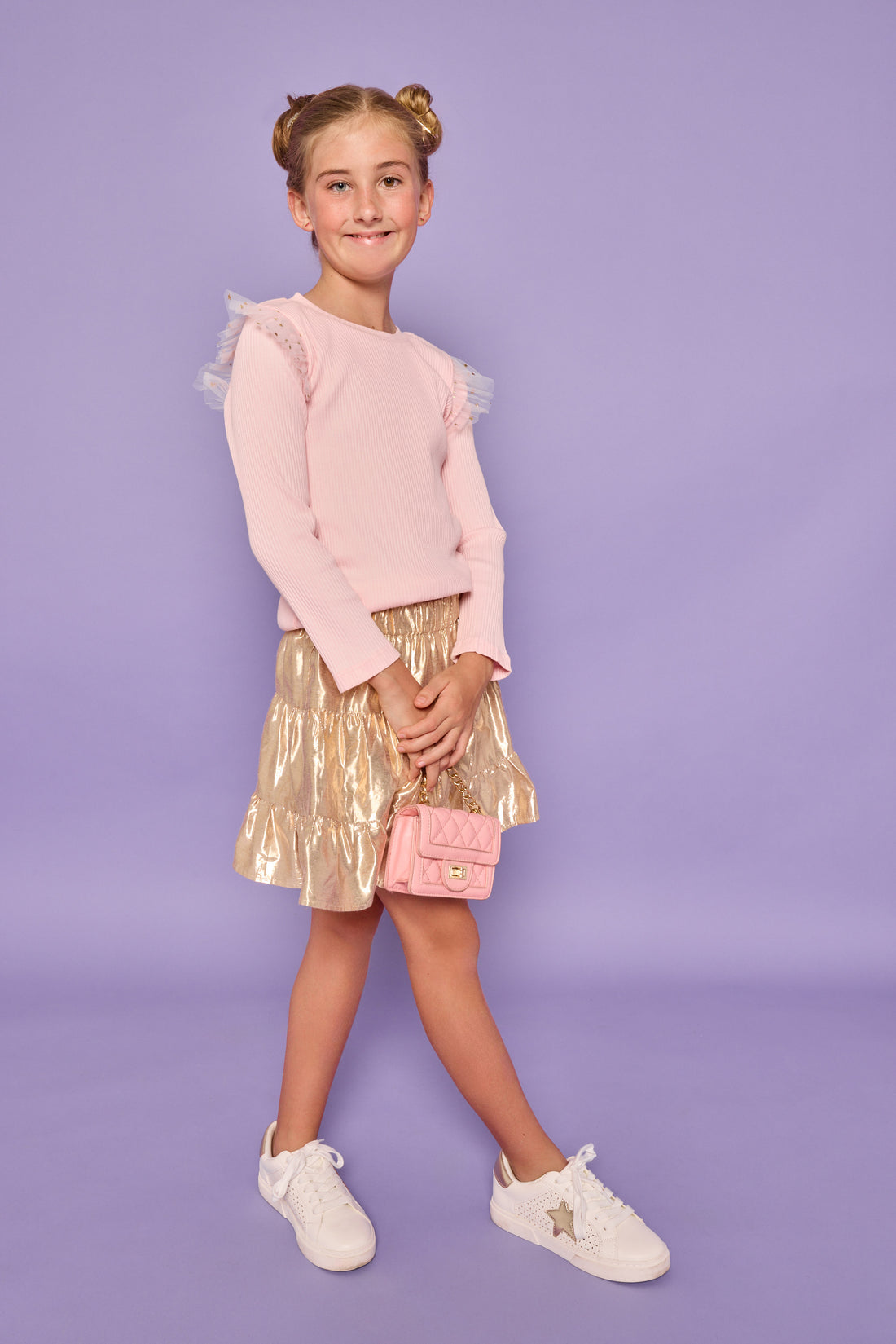 Model can be seen wearing a bubblegum pink ribbed knit long sleeve t-shirt with tulle sparkle star fabric at top of sleeves for elevated look. Paired back with a gold skort in a metallic lurex material that has built in shorts for a secure fit. 