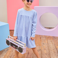 Model can be seen wearing long sleeve velour dress in periwinkle blue. Dress features button closure at back, and pleating detail across bust and bottom tier of dress for an elevated look. 