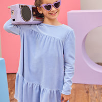 Model can be seen wearing long sleeve velour dress in periwinkle blue. Dress features button closure at back, and pleating detail across bust and bottom tier of dress for an elevated look. 