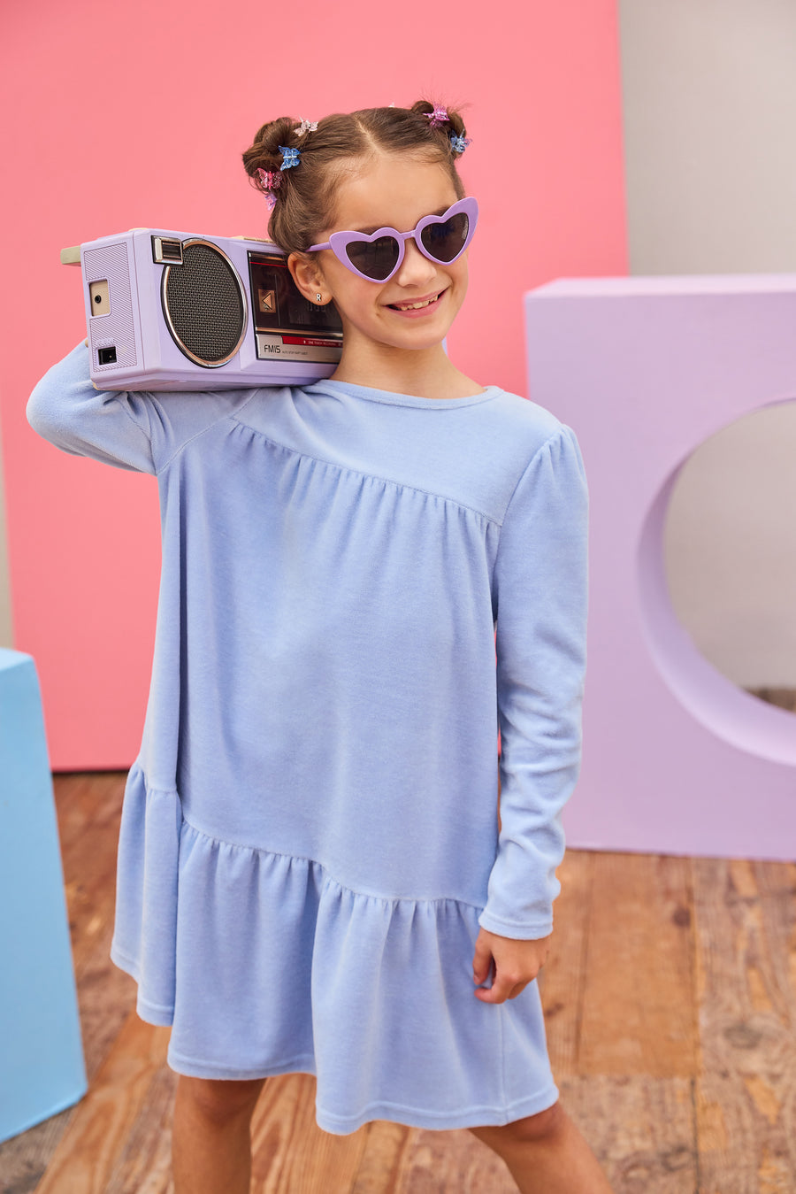 Model can be seen wearing long sleeve velour dress in periwinkle blue. Dress features button closure at back, and pleating detail across bust and bottom tier of dress for an elevated look. 