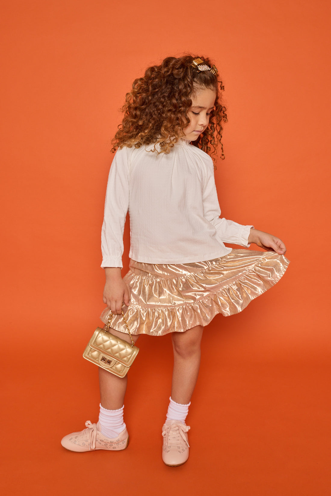 Model can be seen wearing knit pointelle white top with an elastic high neckline  and elastic cuffs paired back with a gold tiered lurex skirt with built in shorts. 