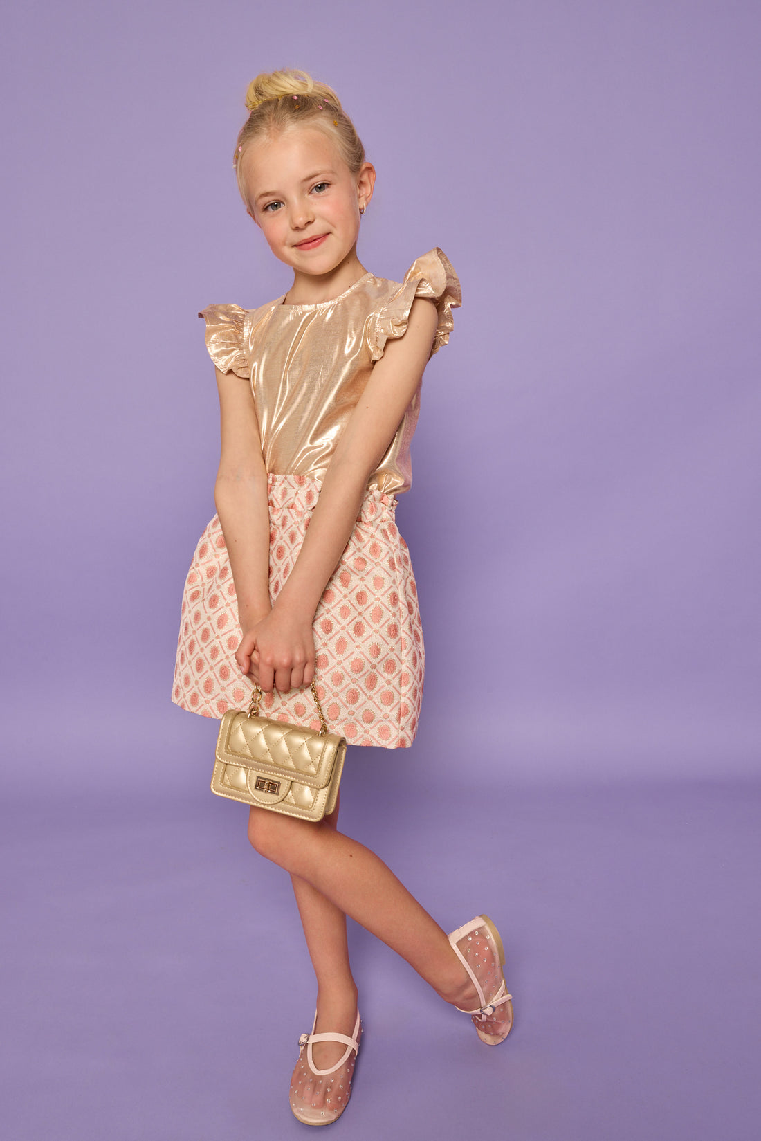 Model can be seen wearing gold metallic lurex top that features angel sleeve detail and button closure at back. Paired back with jacquard skirt with pink and gold metallic details.