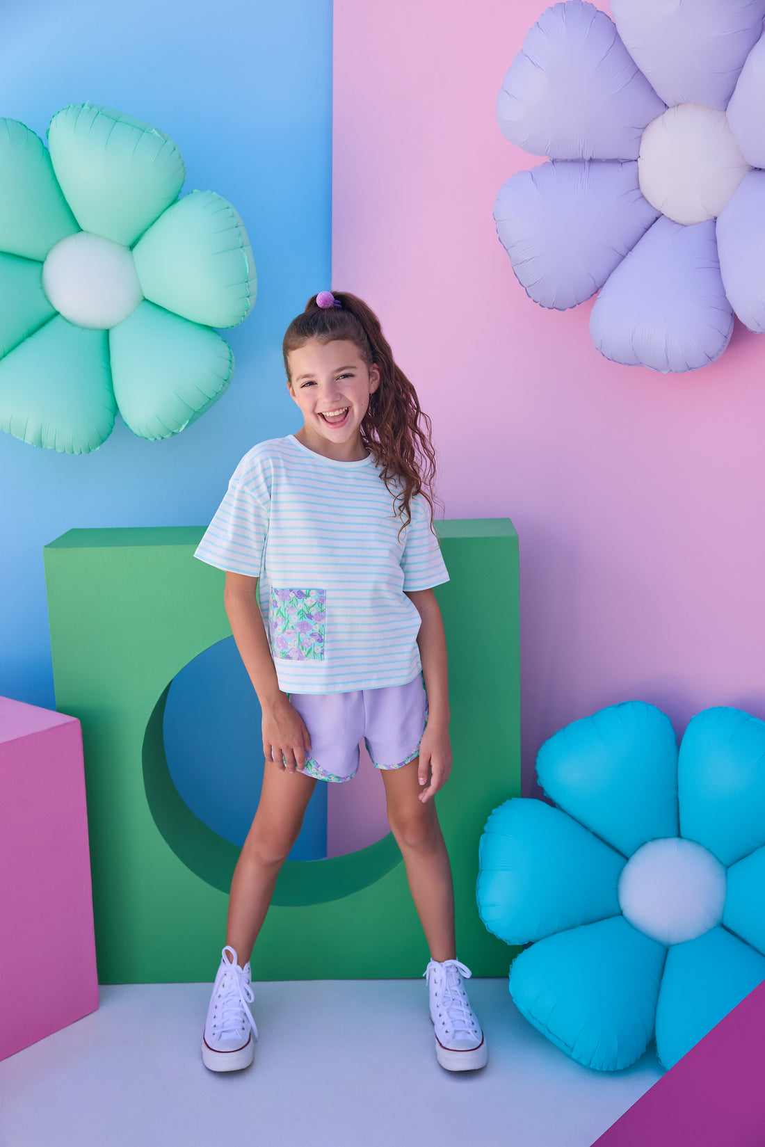 Model can be seen wearing purple knit shorts that feature an elastic waistband and woven purple/blue/green floral trim detail along bottom and sides of shorts to elevate the look. Shorts pair back perfectly with aqua/white knit striped t-shirt that has a pocket detail in the same floral print to pair back with the shorts. 