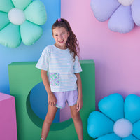Model can be seen wearing purple knit shorts that feature an elastic waistband and woven purple/blue/green floral trim detail along bottom and sides of shorts to elevate the look. Shorts pair back perfectly with aqua/white knit striped t-shirt that has a pocket detail in the same floral print to pair back with the shorts. 