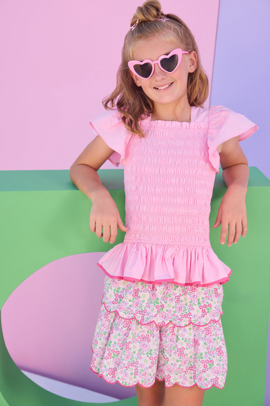 Model can be seen wearing a two tiered skort in a light pink/hot pink/and green floral print. Skort features build in shorts, an elastic waistband, and hot pink embroidered scallop edges for an elevated look. Skort pairs back perfectly with smocked woven short sleeve pink top with hot pink trim to match back perfectly with skort. 