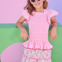 Model can be seen wearing a two tiered skort in a light pink/hot pink/and green floral print. Skort features build in shorts, an elastic waistband, and hot pink embroidered scallop edges for an elevated look. Skort pairs back perfectly with smocked woven short sleeve pink top with hot pink trim to match back perfectly with skort. 