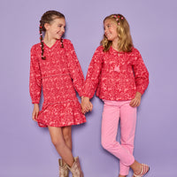 Models can be seen wearing a red and pink floral print in either a woven ruched dress on the left or a woven long sleeve top on the right. The top features tie tassels in the front of the v-neckline for an elevated look, which pairs back perfectly with our corduroy pants in the matching pink color. 