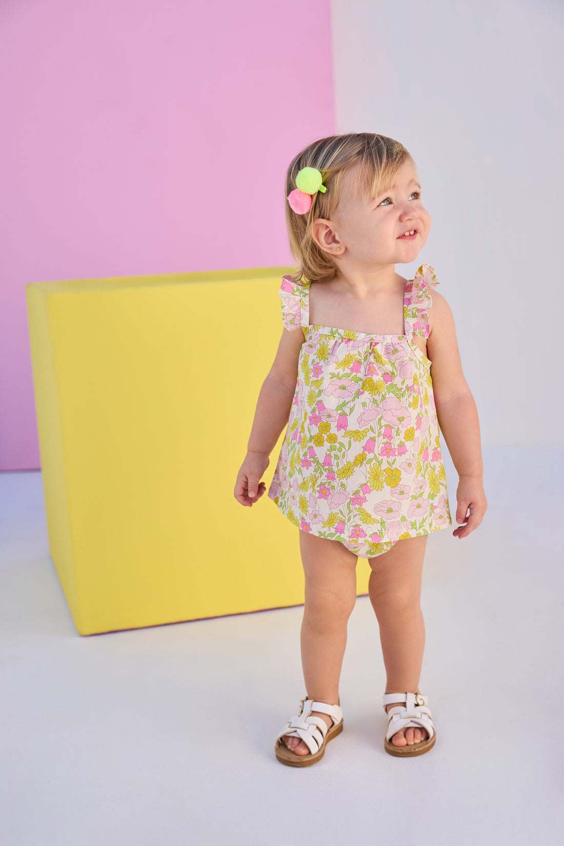 Baby model can be seen wearing a two piece woven bloomer set in a beautiful pink and yellow floral print. Top features button closure at end of straps along back, and bottoms are easy pull on style for a secure but comfy fit. 