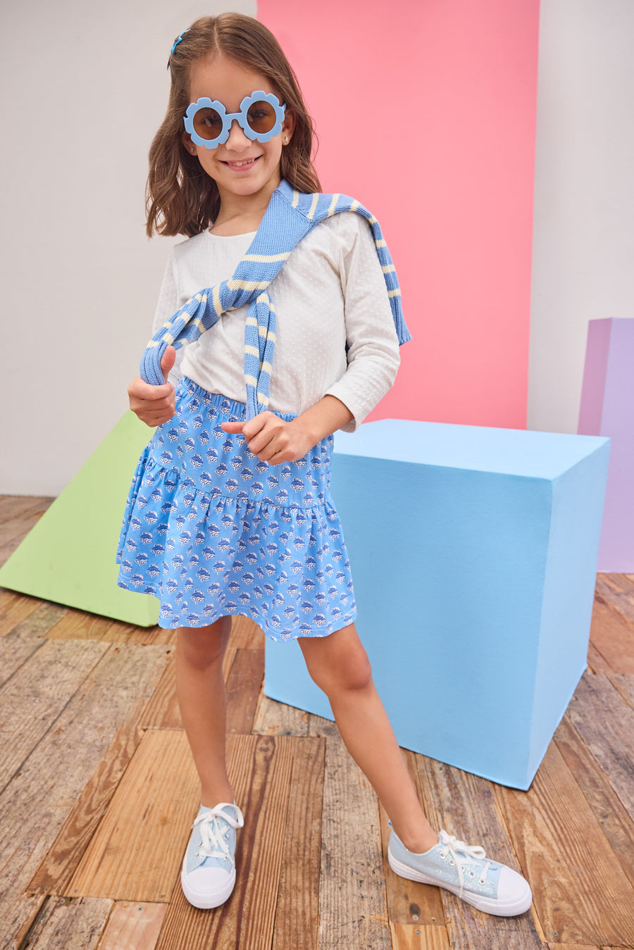 Young girl in a blue and white outfit with sunglasses standing on a wooden floor.