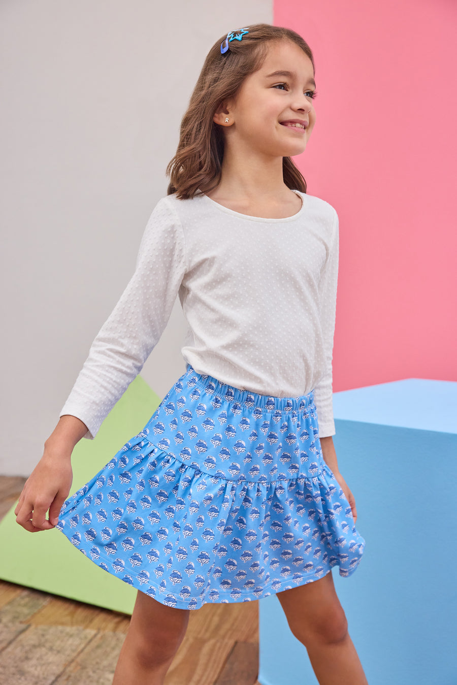 Young girl wearing a white long-sleeve shirt and blue skirt with a pattern against a colorful background