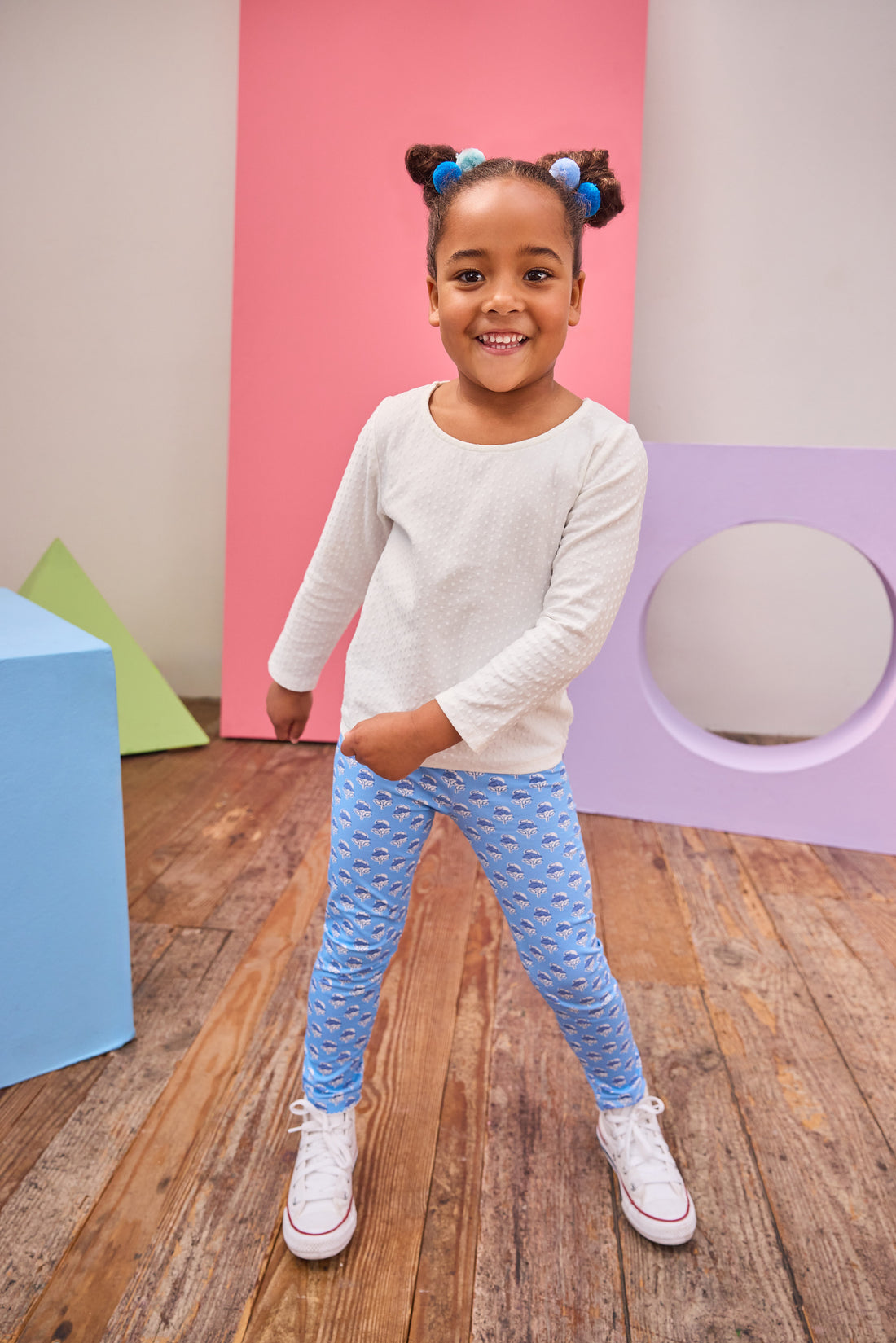 Young girl in a white top and blue patterned pants standing in a room with colorful geometric shapes on the wall.