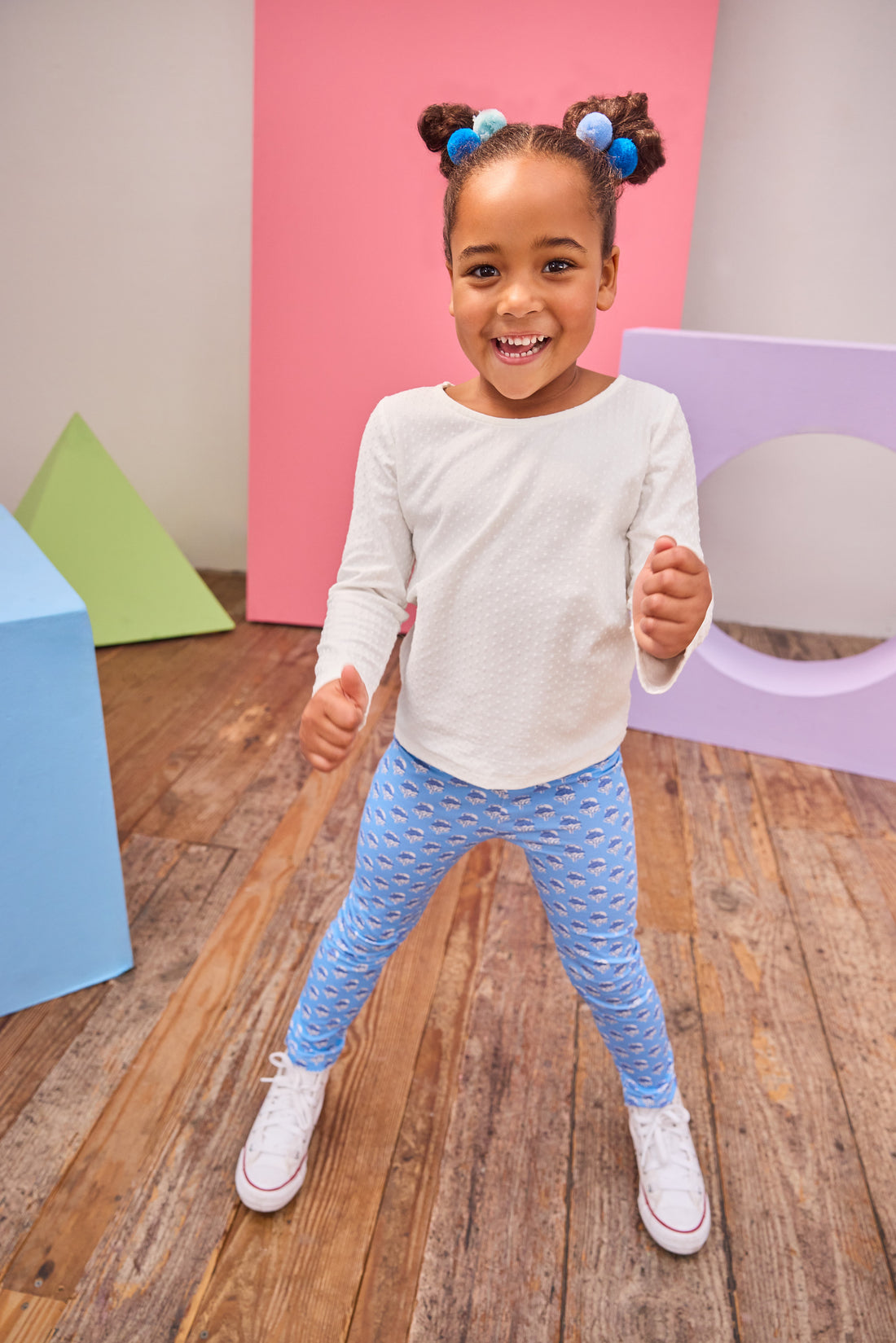 Child wearing a white long-sleeve shirt and blue patterned leggings standing on a wooden floor with colorful geometric shapes in the background.