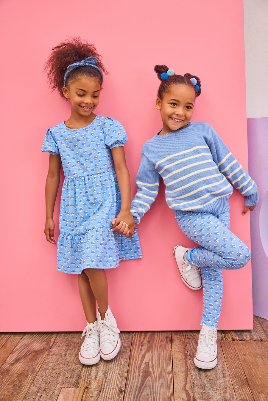 Two young girls in matching blue outfits standing against a pink wall.