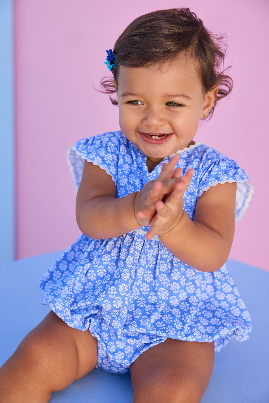 Baby model can be seen wearing a knit bubble in a blue and white floral print. Bubble features white ric rac details on edge of angel sleeves, button closure along back, and stride for an easy put on/pull off access. 