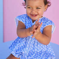 Baby model can be seen wearing a knit bubble in a blue and white floral print. Bubble features white ric rac details on edge of angel sleeves, button closure along back, and stride for an easy put on/pull off access. 