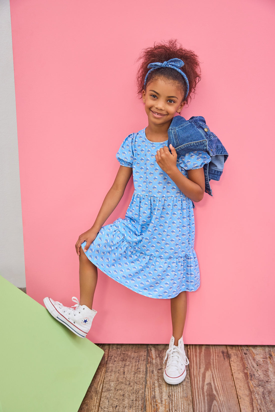 Young girl in a blue dress with a denim jacket against a pink background