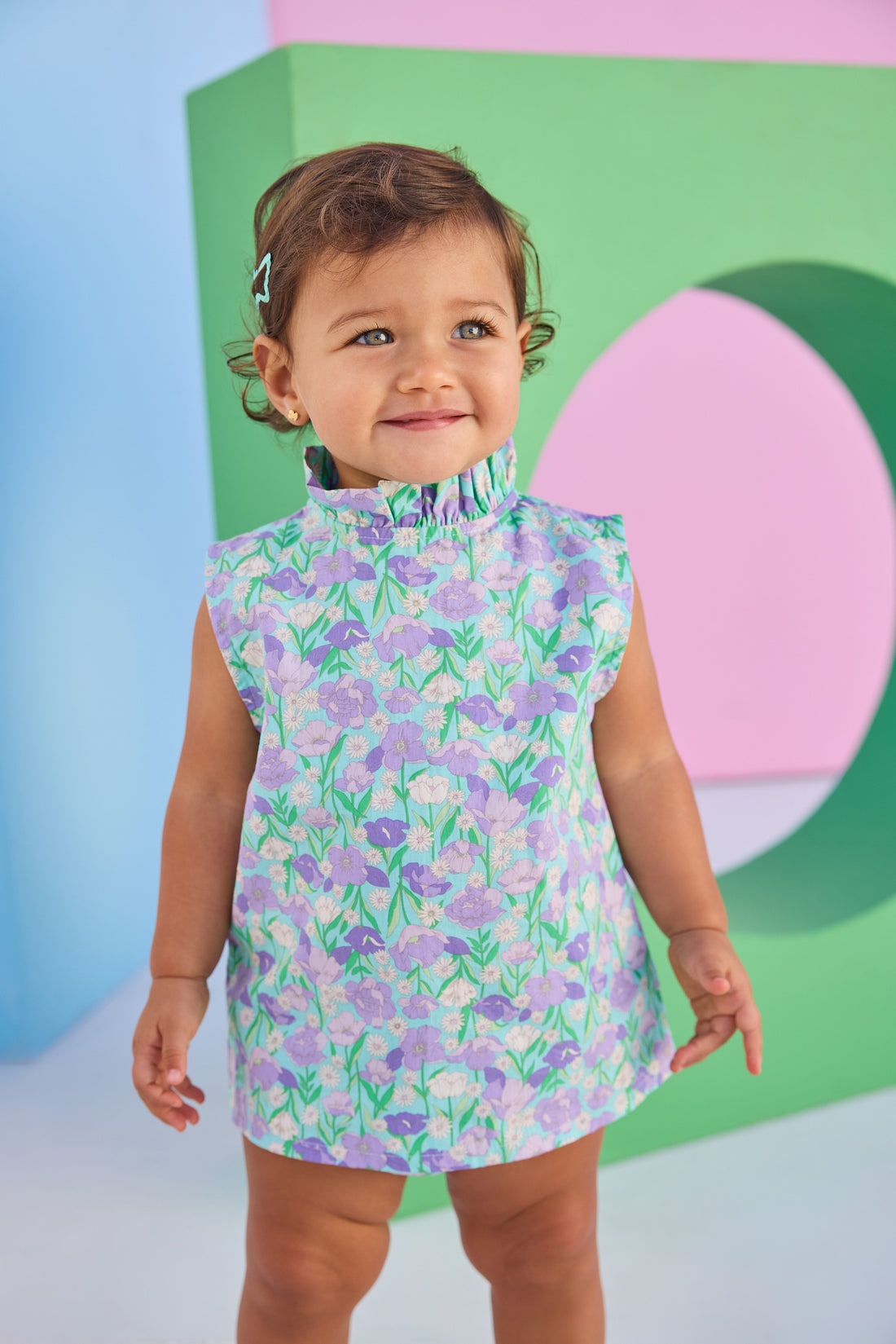 Baby model can be seen wearing a woven bloomer set in an aqua/green/purple floral print. Top of set features button closure at back and bottom features elastic waistband for easy put on/pull off access.