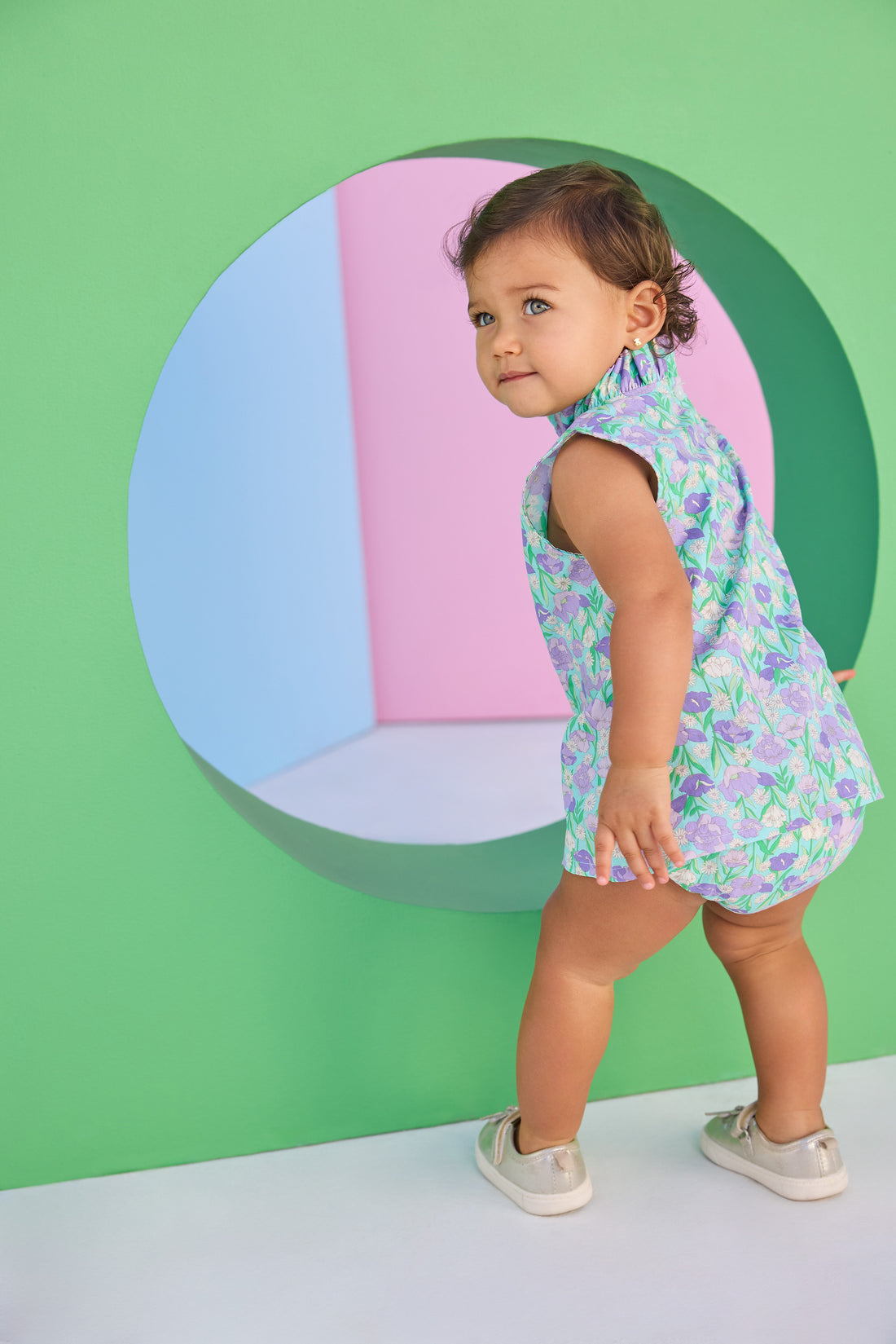 Baby model can be seen wearing a woven bloomer set in an aqua/green/purple floral print. Top of set features button closure at back and bottom features elastic waistband for easy put on/pull off access.