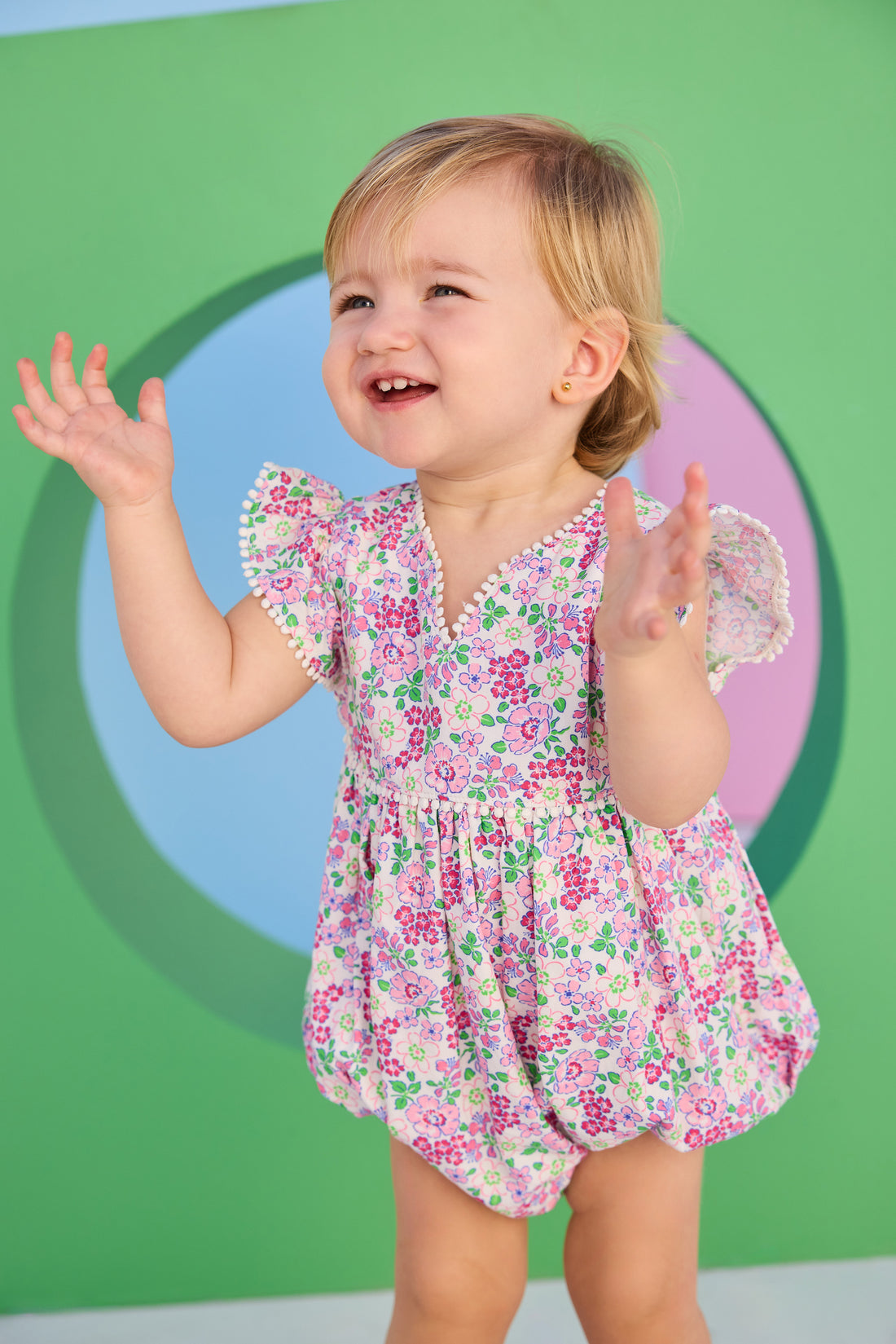 Baby model can be seen wearing a woven bubble in a pink and green floral print. Bubble features white pom pom details on edge of angel sleeves, button closure along back, and stride for an easy put on/pull off access. 
