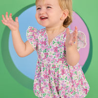 Baby model can be seen wearing a woven bubble in a pink and green floral print. Bubble features white pom pom details on edge of angel sleeves, button closure along back, and stride for an easy put on/pull off access. 