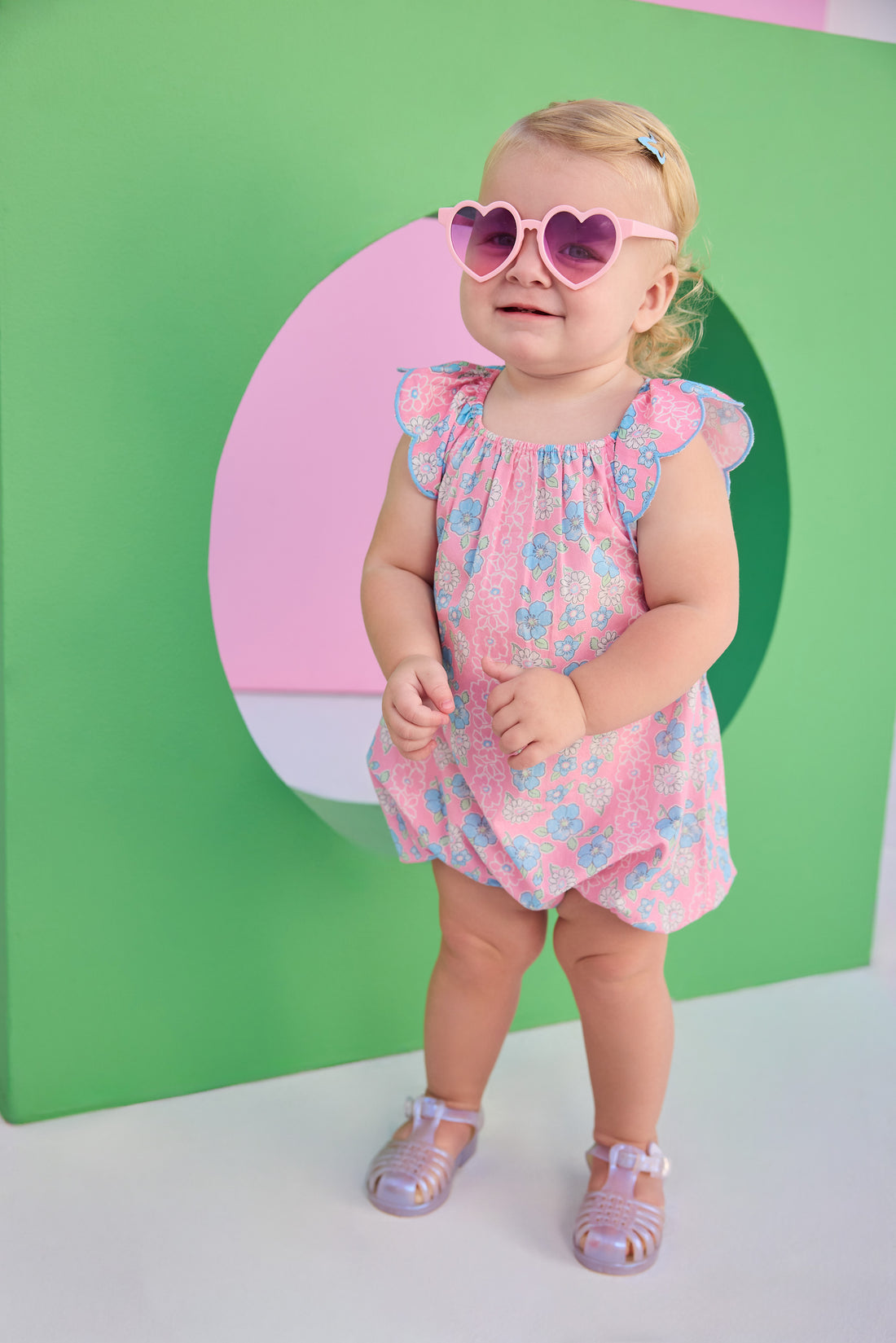 Baby girl seen in a woven pink and blue floral bubble. Bubble features scallop sleeves, elastic bust line, and button snap closure at stride.