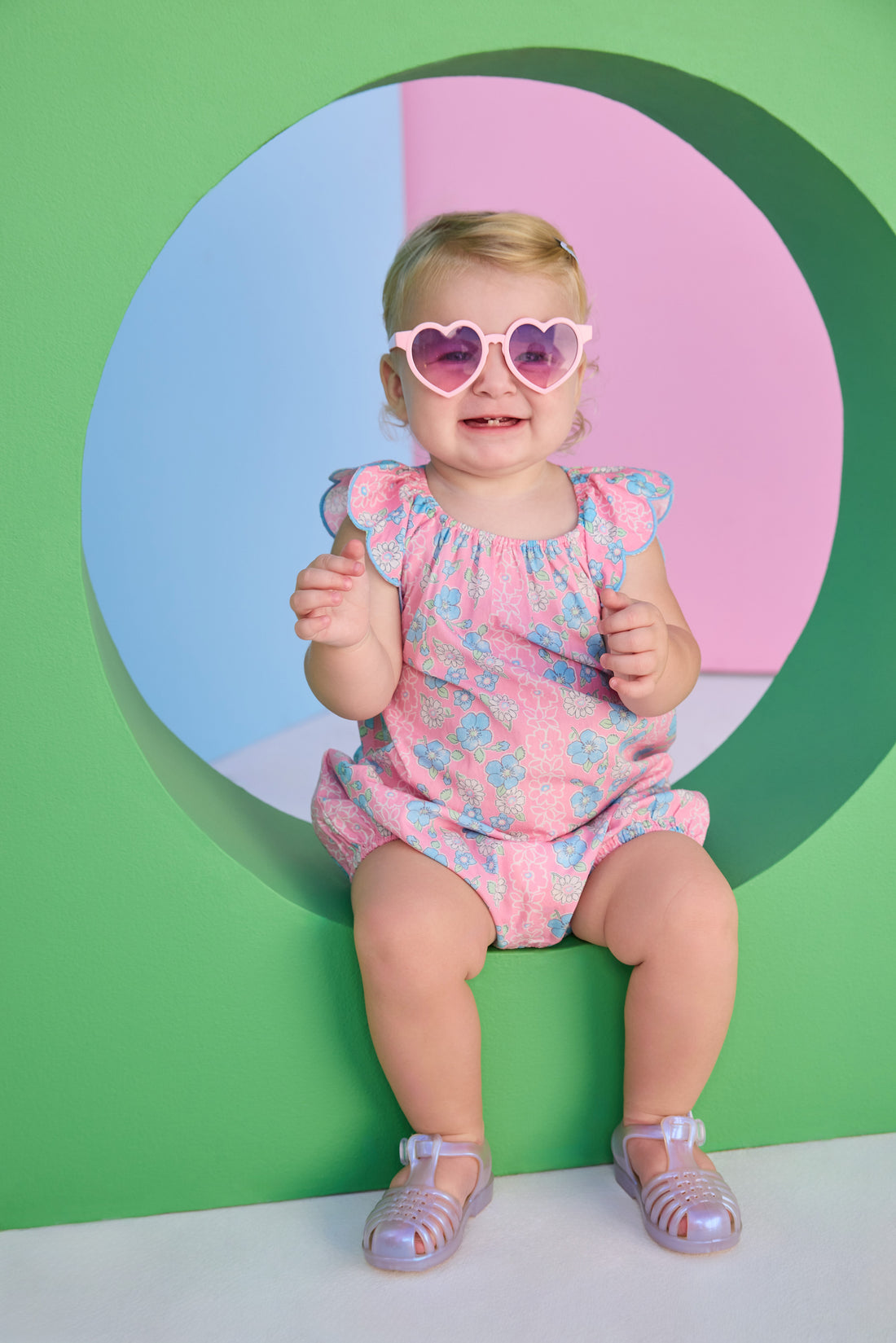 Baby girl seen in a woven pink and blue floral bubble. Bubble features scallop sleeves, elastic bust line, and button snap closure at stride.