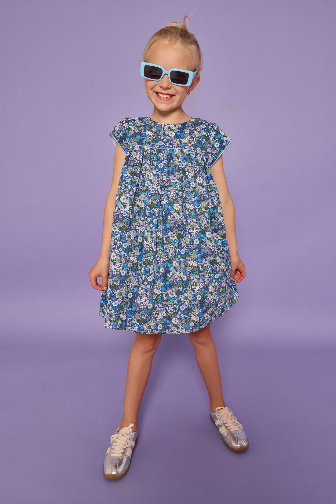 Model can be seen wearing a woven short sleeve dress in a purple/gree/blue floral print. Dress features light blue edge at end of sleeves along with across the bust for an elevated look. Dress also features button closures at back. 