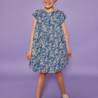 Model can be seen wearing a woven short sleeve dress in a purple/gree/blue floral print. Dress features light blue edge at end of sleeves along with across the bust for an elevated look. Dress also features button closures at back. 