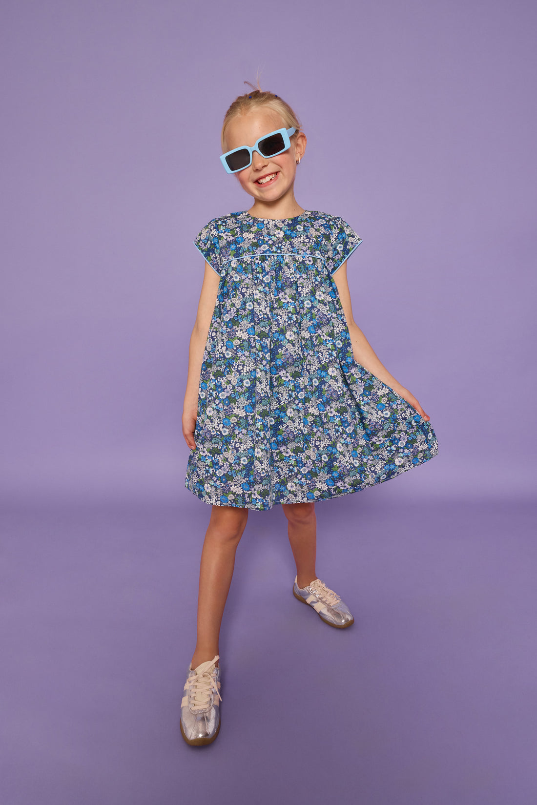 Model can be seen wearing a woven short sleeve dress in a purple/gree/blue floral print. Dress features light blue edge at end of sleeves along with across the bust for an elevated look. Dress also features button closures at back. 