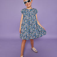 Model can be seen wearing a woven short sleeve dress in a purple/gree/blue floral print. Dress features light blue edge at end of sleeves along with across the bust for an elevated look. Dress also features button closures at back. 