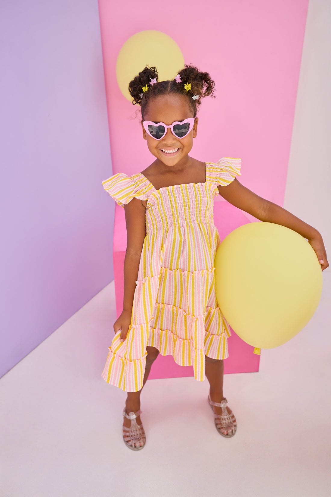 Model can be seen wearing a woven dress in a yellow/pink seersucker fabric. Dress features smocking detail across bust and slight angel sleeves to elevate the look. 