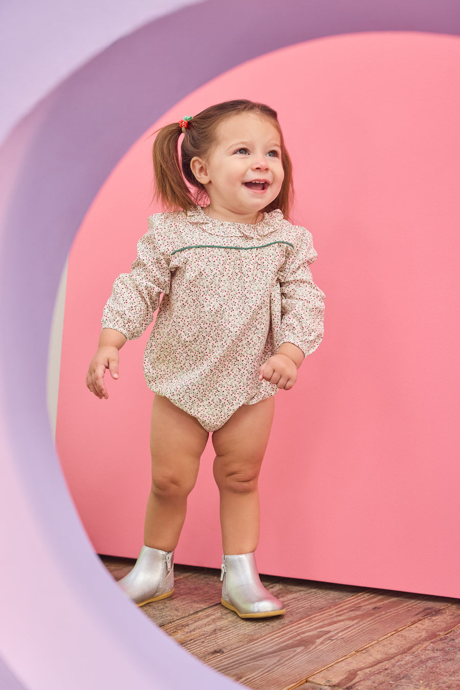Baby girl can be seen wearing a woven bubble in a gorgeous red and green color floral that features a ruffle neckline and snap closure for easy put on/pull off access. 