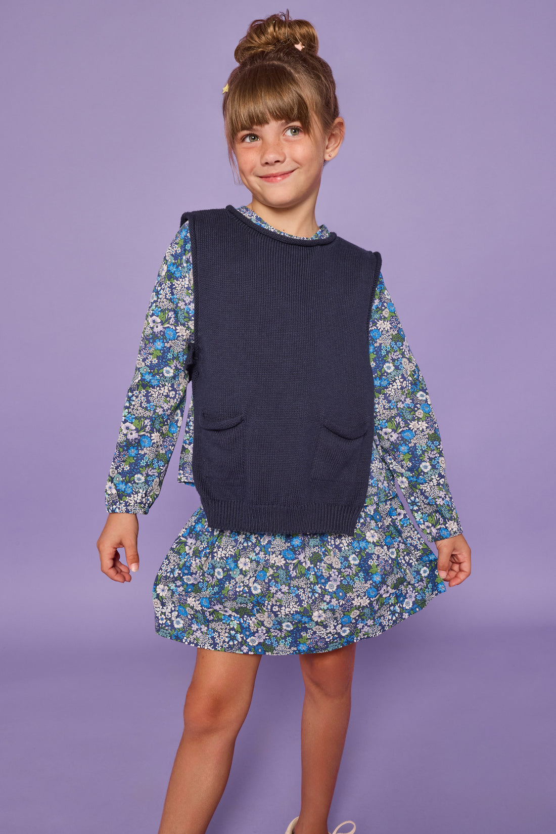 Model can be seen wearing a long sleeve woven dress in a blue/purple/white floral print. Top features ruching across the bust and elastic at the sleeves. Can also be paired underneath a navy sweater vest for a more elevated look.