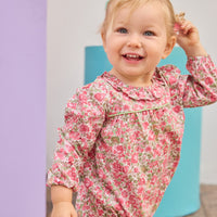 Baby girl can be seen wearing a pink/green floral woven bubble that features  a stunning ruffled collar and ¾ length sleeves. The bubble also has snap closures at the stride, and buttons on back for easy pull on/take off access. 