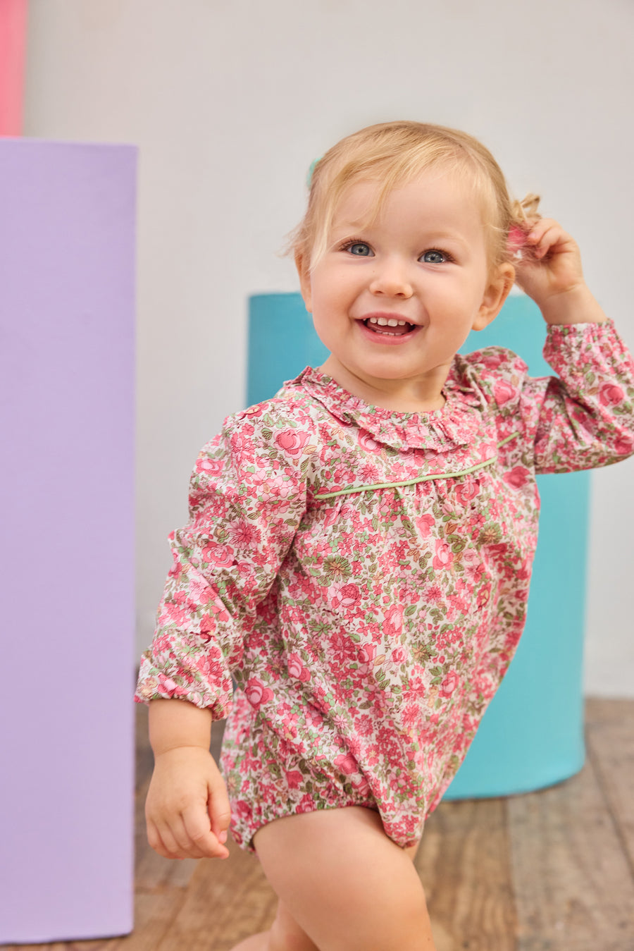 Baby girl can be seen wearing a pink/green floral woven bubble that features  a stunning ruffled collar and ¾ length sleeves. The bubble also has snap closures at the stride, and buttons on back for easy pull on/take off access. 