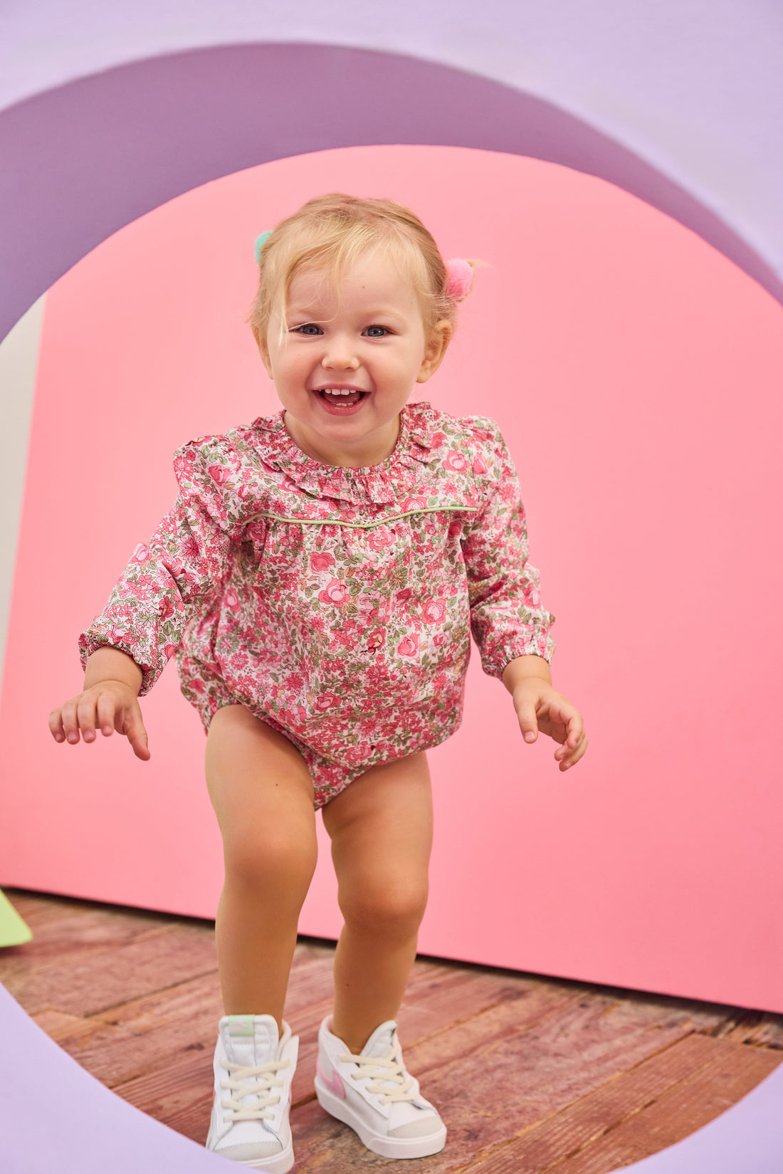 Baby girl can be seen wearing a pink/green floral woven bubble that features  a stunning ruffled collar and ¾ length sleeves. The bubble also has snap closures at the stride, and buttons on back for easy pull on/take off access. 