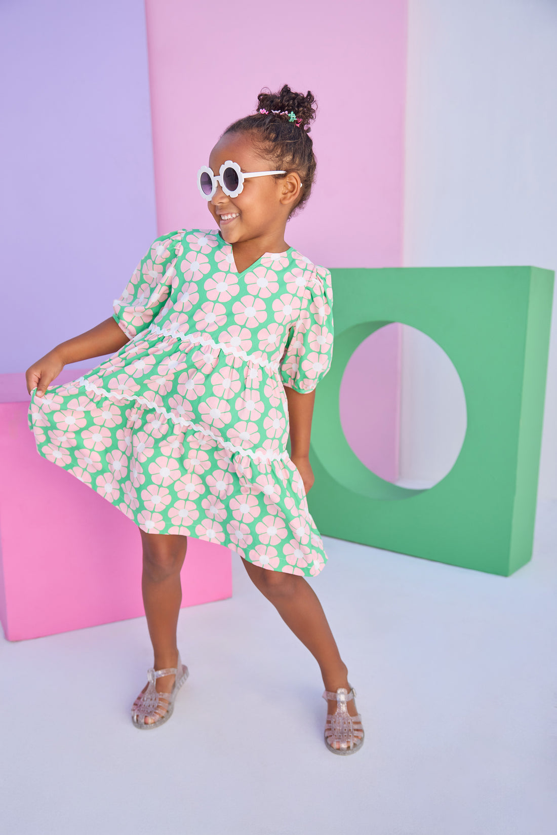 Model can be seen wearing a woven 3/4 sleeve length dress that has button closure along back in a green and salmon pink floral print. Dress features a white ric rac detail on the top and bottom tier of the dress to elevate the look. 