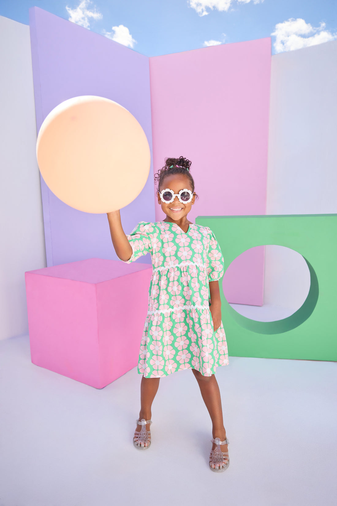 Model can be seen wearing a woven 3/4 sleeve length dress that has button closure along back in a green and salmon pink floral print. Dress features a white ric rac detail on the top and bottom tier of the dress to elevate the look. 