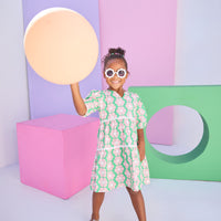 Model can be seen wearing a woven 3/4 sleeve length dress that has button closure along back in a green and salmon pink floral print. Dress features a white ric rac detail on the top and bottom tier of the dress to elevate the look. 