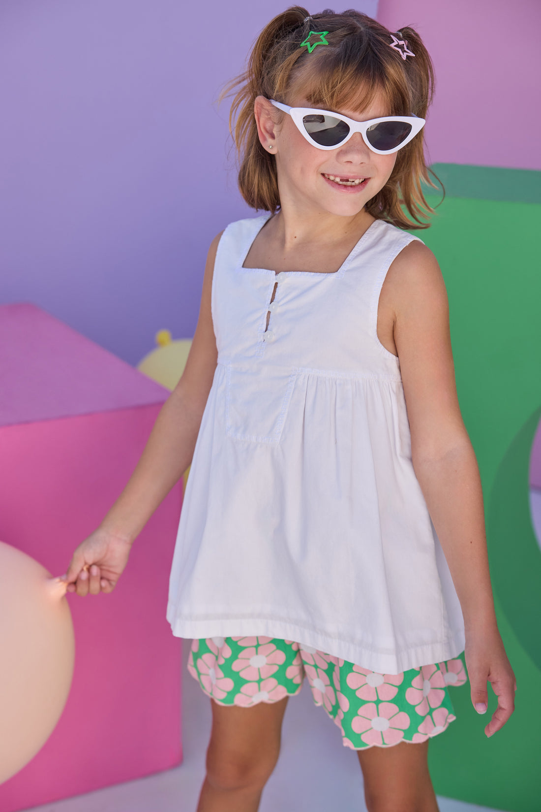 Model can be seen wearing shirred shorts in a salmon pink/green floral print. Shorts feature smocking along waist and scallop edges to elevate the look. Shorts pair back perfectly with woven white top with a square neckline for an elevated but comfy look. 