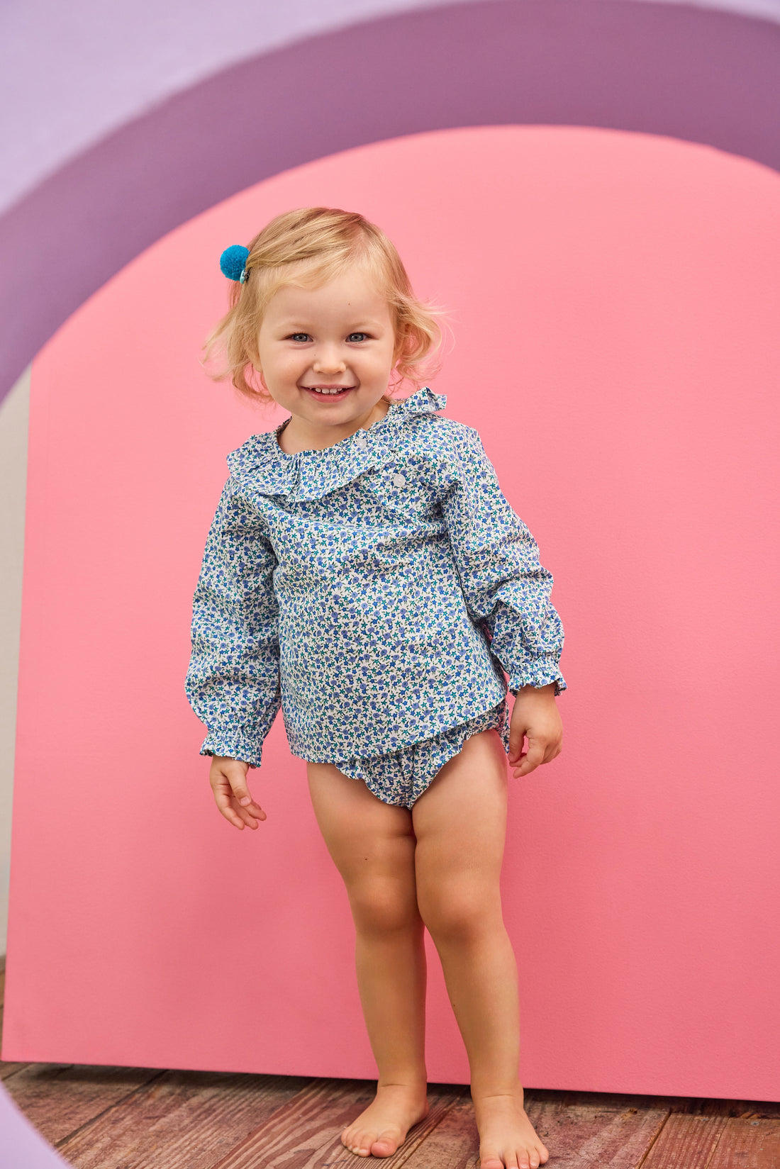 Baby model can be seen wearing a woven bloomer set in a blue floral print that features two  button closure on front of top of the set at neckline and elastic waist on bottoms for easy put on/take off access. 