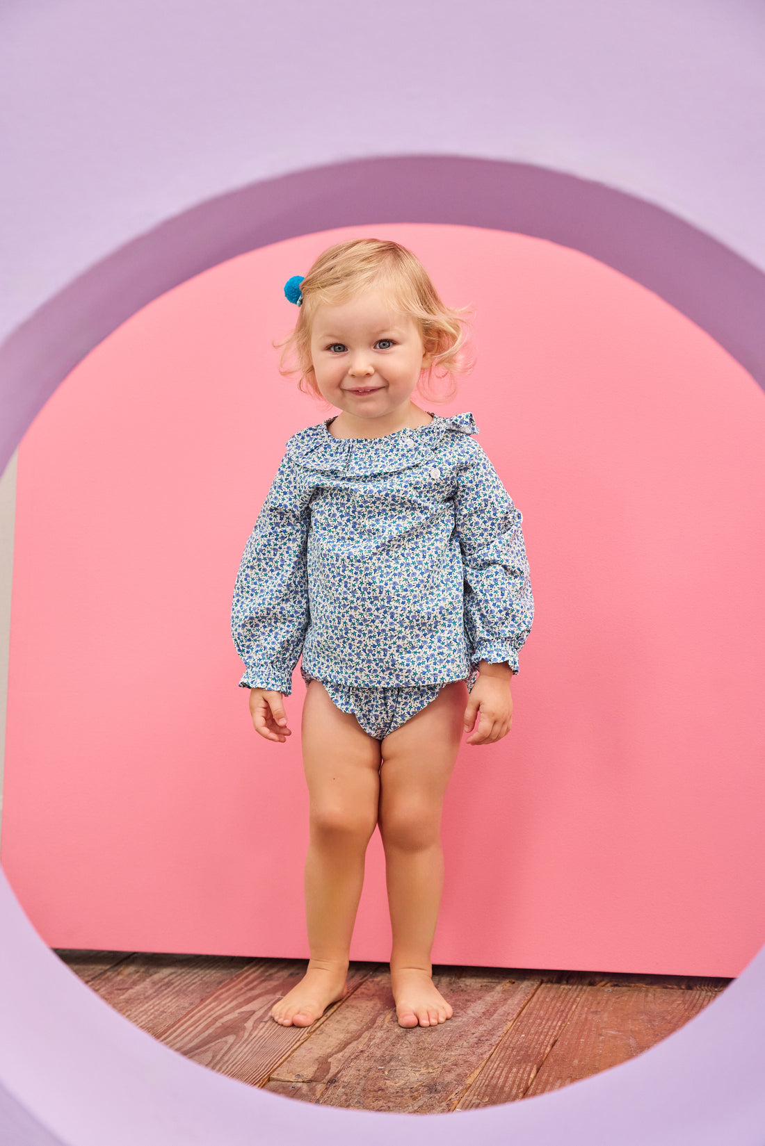 Baby model can be seen wearing a woven bloomer set in a blue floral print that features two  button closure on front of top of the set at neckline and elastic waist on bottoms for easy put on/take off access. 
