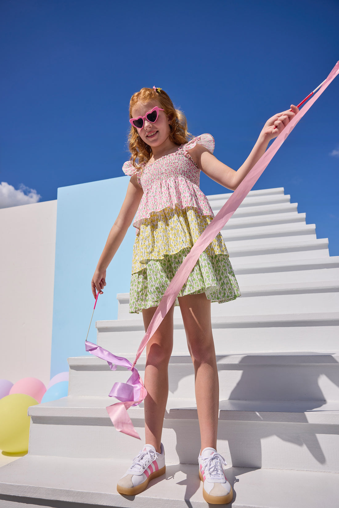 Tween model can be seen wearing a woven three tiered dress that features the same floral print recolored in pink/yellow/and green. Dress features angel sleeve detail on straps and smocking detail across bust for a comfy but secure fit.