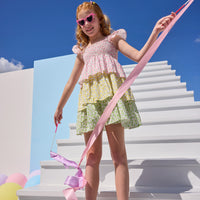 Tween model can be seen wearing a woven three tiered dress that features the same floral print recolored in pink/yellow/and green. Dress features angel sleeve detail on straps and smocking detail across bust for a comfy but secure fit.