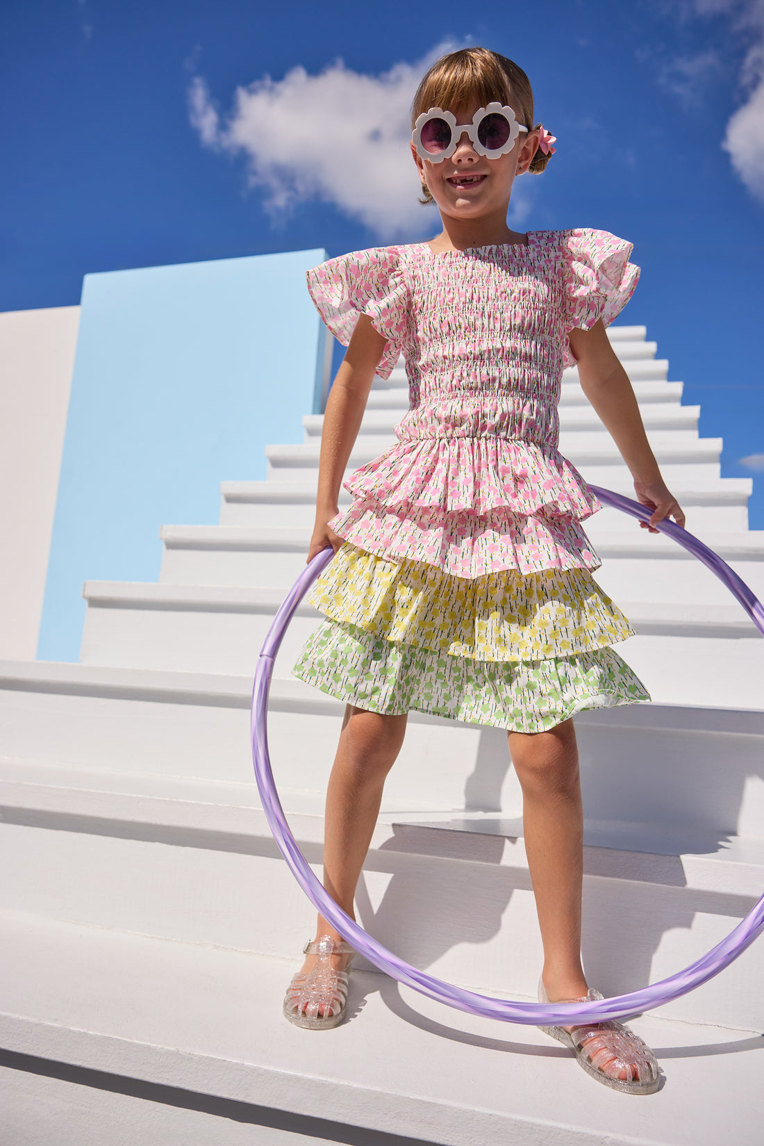 Model can be seen wearing woven pink floral print top that features elastic smocking throughout the body of the top for a stretchy but secure fit. Top pairs back perfectly with three tiered skort in matching floral print in pink/yellow/green floral tiers. 