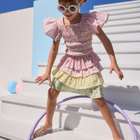 Model can be seen wearing woven pink floral print top that features elastic smocking throughout the body of the top for a stretchy but secure fit. Top pairs back perfectly with three tiered skort in matching floral print in pink/yellow/green floral tiers. 