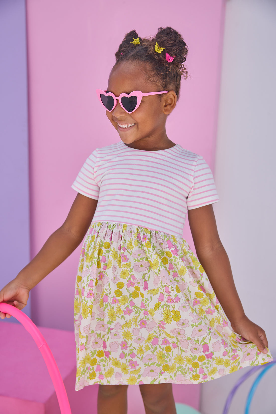 Model can be seen wearing dress in half knit fabric and half woven fabric. The top of the dress is a knit pink and white fabric and the bottom of the dress is a woven pink and yellow floral fabric. Dress features button closure at back as well for a secure but comfy fit.