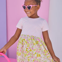 Model can be seen wearing dress in half knit fabric and half woven fabric. The top of the dress is a knit pink and white fabric and the bottom of the dress is a woven pink and yellow floral fabric. Dress features button closure at back as well for a secure but comfy fit.