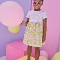 Model can be seen wearing dress in half knit fabric and half woven fabric. The top of the dress is a knit pink and white fabric and the bottom of the dress is a woven pink and yellow floral fabric. Dress features button closure at back as well for a secure but comfy fit.