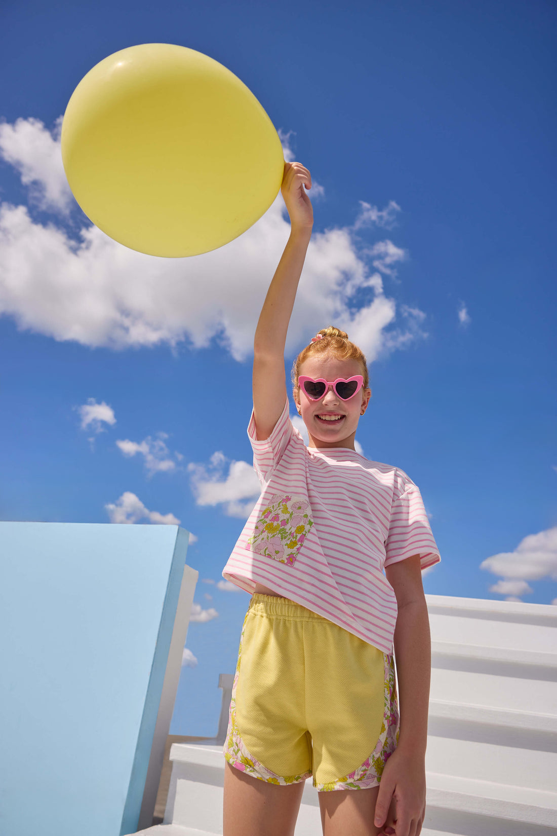 Model can be seen wearing yellow knit shorts that feature an elastic waistband and woven pink/yellow floral trim detail along bottom and sides of shorts to elevate the look. Shorts pair back perfectly with pink/white knit striped t-shirt that has a pocket detail in the same floral print to pair back with the shorts. 